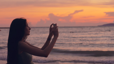 Young tourist woman photographs ocean view with smartphone during sunset at beachの写真素材