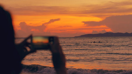 Closeup of young tourist woman photographs ocean view with smartphone during sunset at beachの写真素材