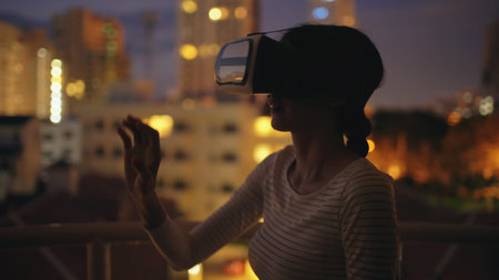 Young woman on rooftop terrace using virtual reality headset and having VR experience at night