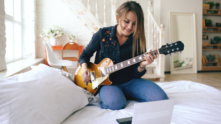 Attractive young girl learning to play electric guitar with notebook sit on bed in bedroom at homeの写真素材