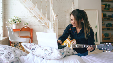 Attractive young girl learning to play electric guitar with notebook sit on bed in bedroom at homeの写真素材