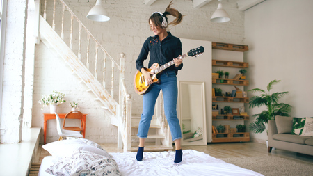 Funny young woman in headphones jumping on bed with electric guitar in bedroom at home indoorsの写真素材