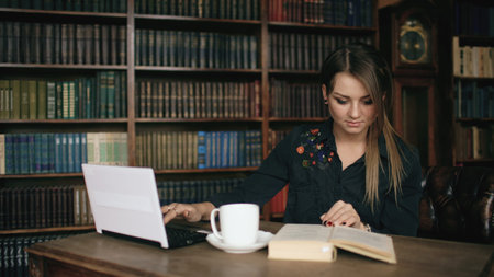 Smiling student girl working on laptop and read book in university library indoors