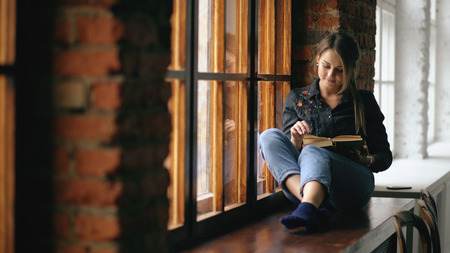 beautiful young student girl reading book sit on windowsill in university classroom indoorsの写真素材