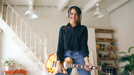 Beautiful young smiling girl in headphones posing with electric guitar at home studio indoorsの写真素材