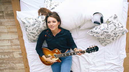 Attractive young girl learning to play electric guitar lie on bed in bedroom at home indoorsの写真素材
