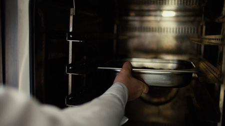 Closeup of professional chef man put meat dish into oven at restaurant kitchenの写真素材