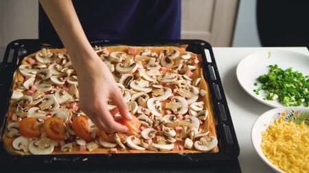 Young beautiful woman cook put tomatos on pizza dough in the kitchen at homeの写真素材