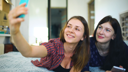 Happy cheerful sisters making selfie in morning and have fun on bed in bedroom at homeの写真素材