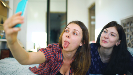 Happy cheerful sisters making selfie in morning and have fun on bed in bedroom at homeの写真素材