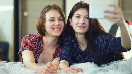 Two happy women friends lying in bed and making selfie in morning and have fun on bedの写真素材