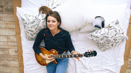 Attractive young girl learning to play electric guitar lie on bed in bedroom at home indoorsの写真素材