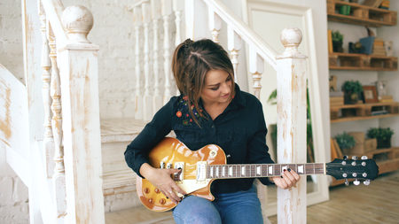 Attractive young girl learning to play electric guitar sit on stairs in bedroom at homeの写真素材