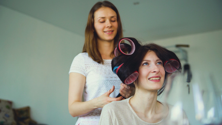 Two happy women friends make fun curler hairstyle each other and have fun at homeの写真素材
