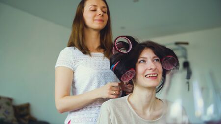 Two happy women friends make fun curler hairstyle each other and have fun at homeの写真素材