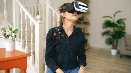 Woman in VR headset looking up and trying to touch objects in virtual reality at home indoorsの写真素材