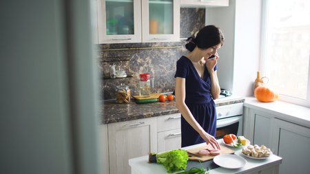 Beautiful girl cook while talking on the mobile phone and smiling in kitchen at homeの写真素材