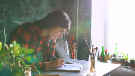 Young woman artist painting sketch on paper notebook with pencil. Bright sun flare from window