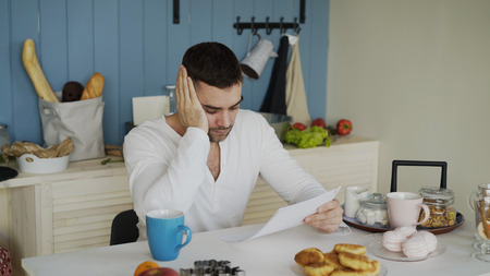 Upset young man reading letter with unpaid bill in the kitchen at homeの写真素材