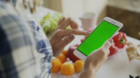 Closeup of womans hand browsing smartphone with green screen on kitchen at homeの写真素材