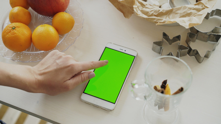 Closeup of womans hand browsing smartphone with green screen on kitchen table at homeの写真素材