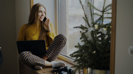 Cheerful woman photographer talking phone and sitting on windowsill with camera and laptop computer...
