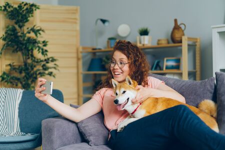 Happy young girl is taking silfie with lovable dog using smartphone camera having fun sitting on couch holding device. Youth and domestic animals concept.の写真素材