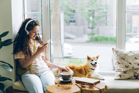 Pretty girl is listening to music with headphones and using smartphone sitting on window sill in cafe with lovely well-bred dog lying near her. Youth and pets concept.の写真素材