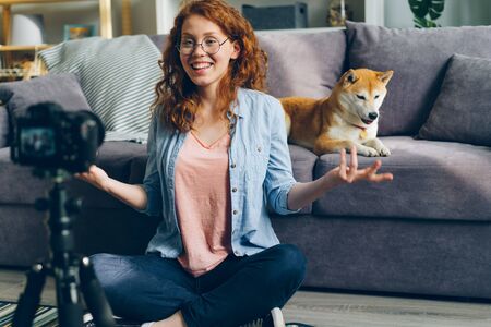 Beautiful girl student recording video with dog for internet vlog at home sitting on floor talking and gesturing using camera on tripod. People and blogging concept.の写真素材
