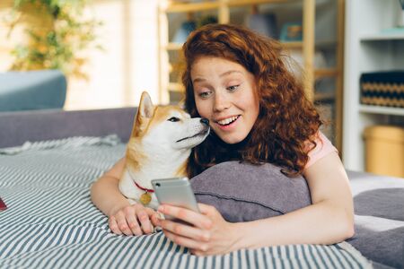 Happy young woman is taking silfie with adorable dog using smartphone camera having fun lying on couch holding device. Youth and domestic animals concept.の写真素材