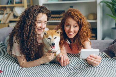 Joyful sisters attractive girls are taking selfie with adorable doggy using smartphone camera lying on couch at home smiling having fun. People and self portrait concept.の写真素材