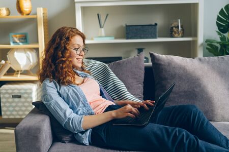 Happy young lady is using laptop typing sitting on sofa in apartment with gadget smiling enjoying social media. People, modern technology and youth concept.の写真素材