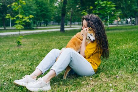 Pretty young woman is hugging shiba inu dog and smiling sitting on grass in park enjoying friendship with animal. People, affection and cute pets concept.の写真素材