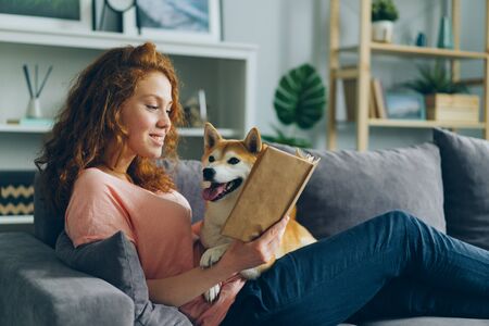 Pretty student young woman is reading book in cozy apartment smiling and petting adorable dog sitting on comfy couch at home. Animals and hobby concept.