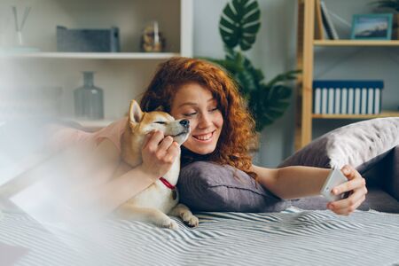 Teenage girl taking selfie with cute puppy using smartphone at home lying on couch in studio apartment smiling posing. People, animals and photographs concept.の写真素材