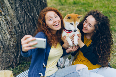 Attractive young ladies taking selfie with small dog in park posing for smartphone camera smiling...