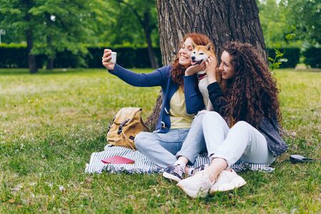Female students joyful girls taking selfie with adorable puppy in park using smartphone camera having fun together. People, youth and animals concept.の写真素材
