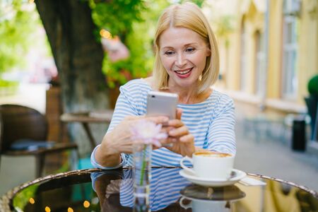 Attractive mature blonde in casual clothing is using smartphone enjoying summer day in open air cafe. Modern technology, people and leisure time concept.の写真素材