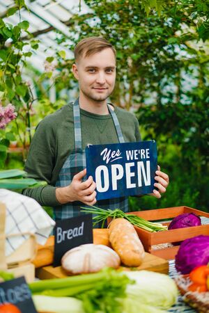 Cheerful farmer is holding we are open sign working in farm market selling organic food inviting people to harvest sale. Small business and shopping concept.の写真素材