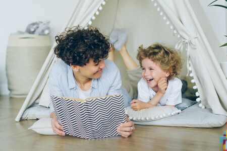 Loving mother is reading interesting book to cute child lying on floor in cosy tent indoors in modern light apartment smiling speaking. People and childhood concept.の写真素材
