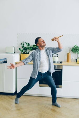 Portrait of silly middle-aged guy having fun in kitchen singing in spoon dancing enjoying life alone in apartment. Modern lifestyle, people and interior concept.の写真素材