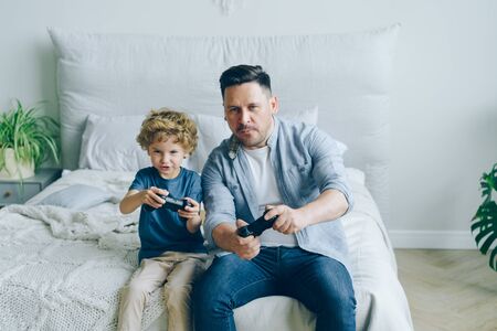 Cute kid is playing video game with happy father holding joysticks having fun in bed at home enjoying gadget and modern entertainment. People and devices concept.の写真素材