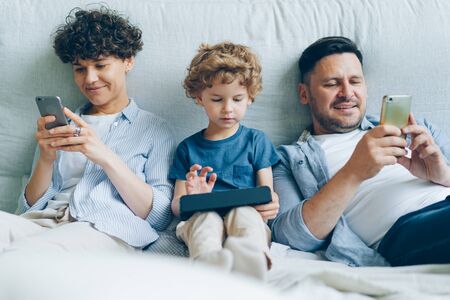 Man and woman are using smartphones checking social media while little kid is playing game on tablet in bed at home. Internet addiction and family concept.の写真素材