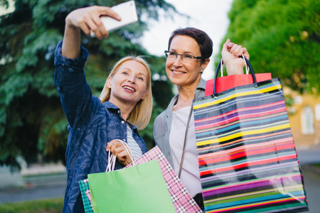 Beautiful mature women friends are taking selfie with smartphone camera posing with bright shopping...