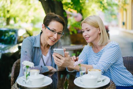 Beautiful mature women friends are using smartphone talking laughing looking at screen sitting in street cafe at table. Modern technology and friendship concept.の写真素材