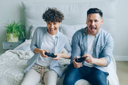 Cute young couple boyfriend and girlfriend are playing videogame holding joysticks sitting on bed in bedroom at home. Emotional people are enjoying computer game.の写真素材