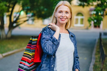 Portrait of good-looking stylish blonde holding shopping bags outdoors in city street smiling looking at camera standing alone on sidewalk. Emotions and consumerism concept.の写真素材
