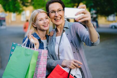 Cheerful people friends taking selfie holding paper bags with purchases outdoors smiling using smartphone camera. People, friendship and shopaholism concept.の写真素材