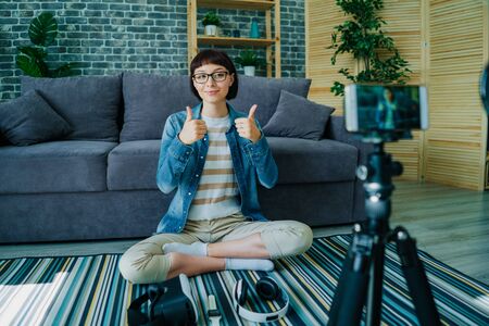 Pretty young lady blogger recording video with smartphone showing thumbs-up sitting on floor at home. Blogging, creative youth and apartment concept.の写真素材