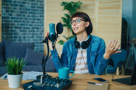 Female blogger recording audio in sound studio talking in microphone with serious face sitting at desk alone. Blogging, business and modern devices concept.の写真素材